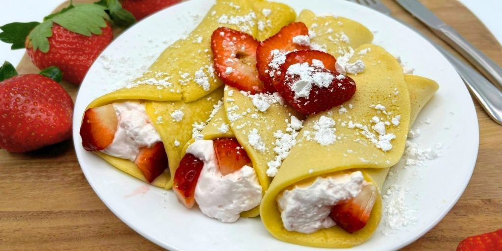 3 rolled up homemade crepes filling with whipped cream and fresh strawberries topped with powdered sugar and strawberry slices
