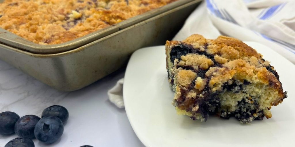 a slice of high altitude blueberry coffee cake with streusel topping sitting on a white plate next to the pan