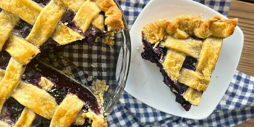 1 slice of high altitude blueberry pie sitting on a while plate next to the whole pie