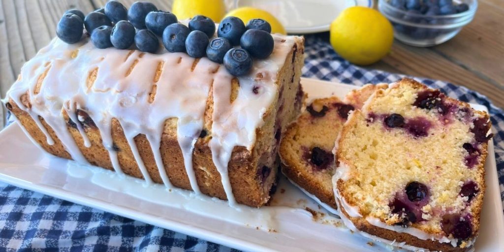 a loaf of high altitude lemon blueberry bread topped with lemon glaze with 2 slices cut off