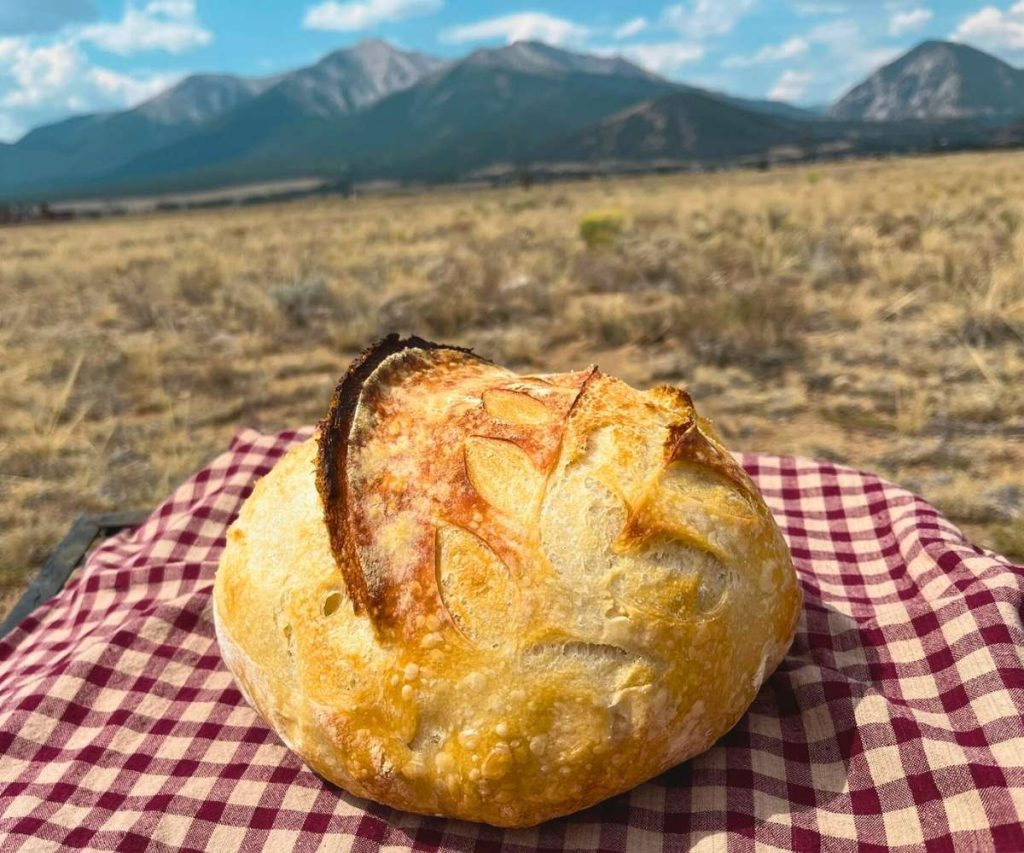 a loaf of high altitude sourdough bread sitting on a red and white tea towel in front of the Colorado Rocky Mountains