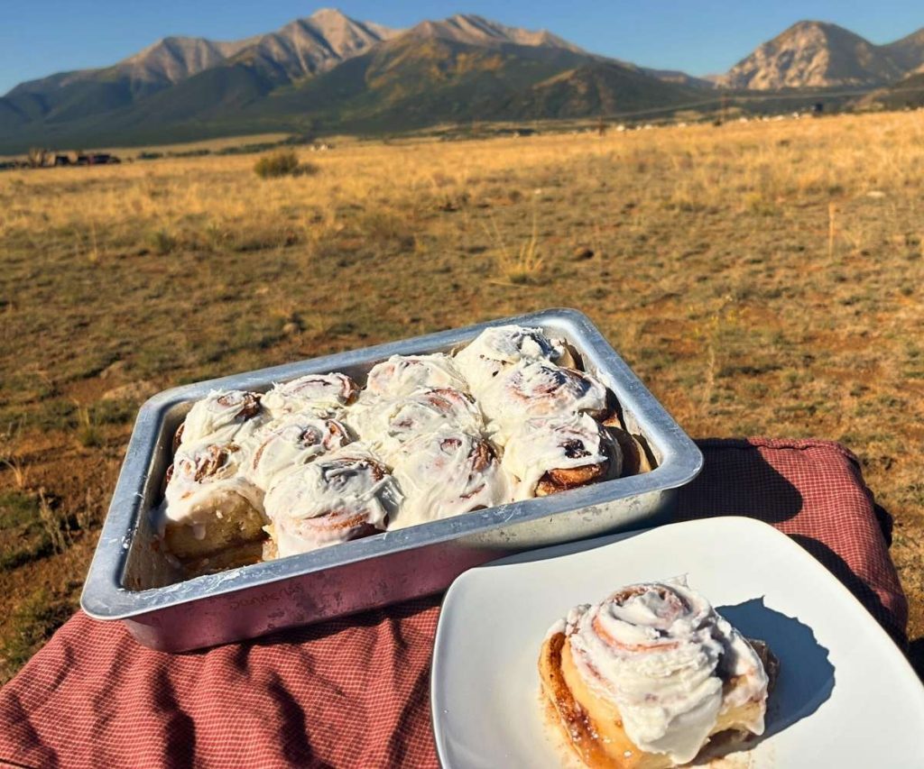 a high altitude sourdough cinnamon roll on a plate in front of a 9x13 pan of rolls in front of the Colorado Rocky Mountains