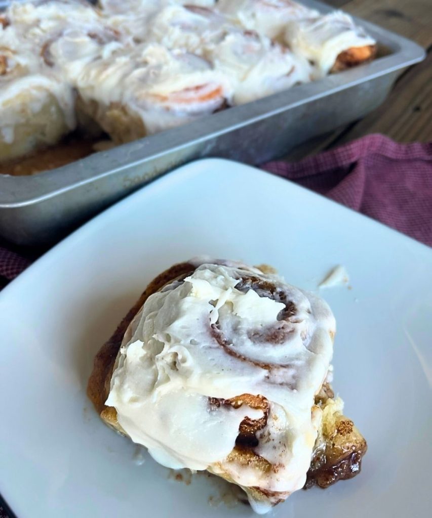 a sourdough cinnamon roll on a plate in front of a 9x13 pan of high altitude sourdough cinnamon rolls